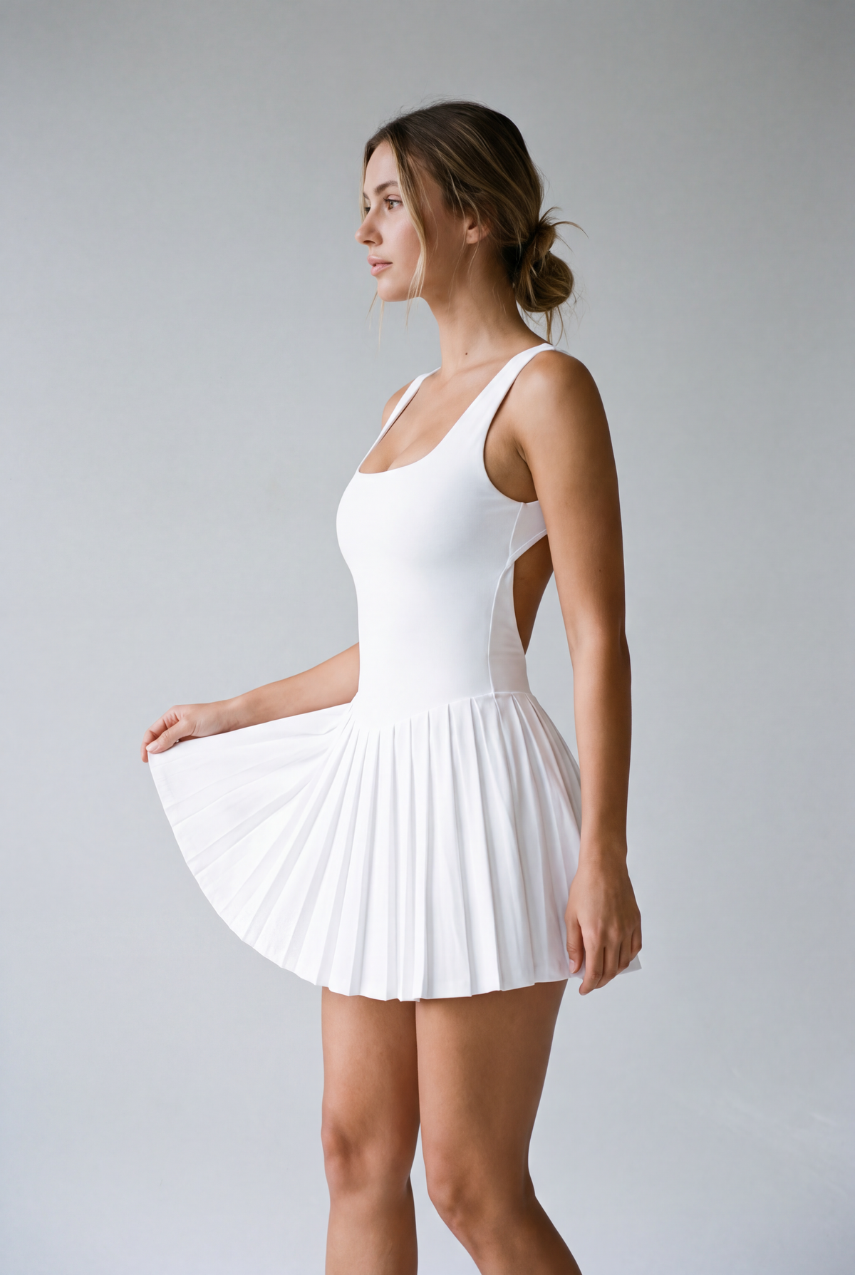A woman modeling a white sleeveless dance or gymnastics leotard dress with a pleated skirt, shown in profile against a light gray studio background.
